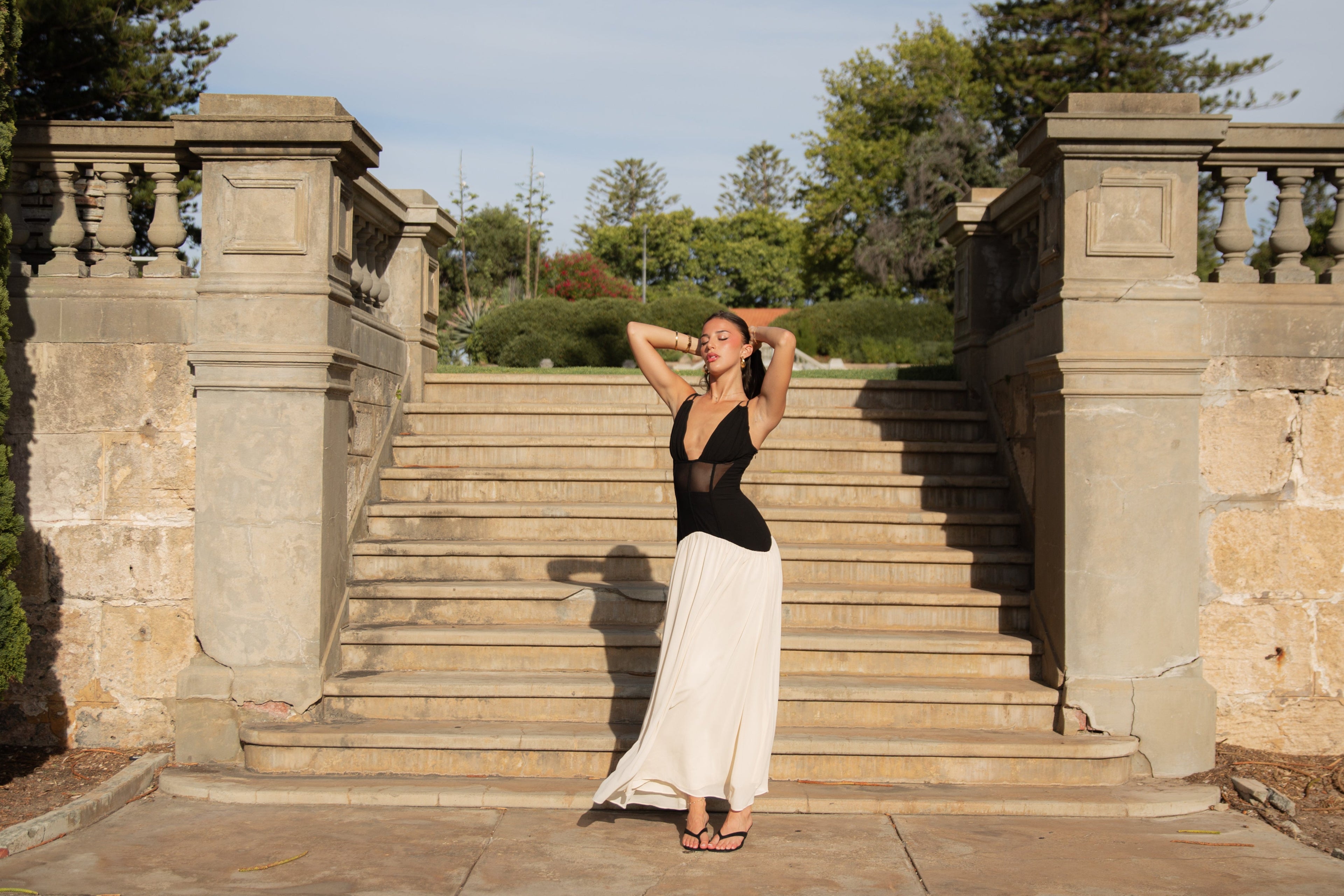 Woman in black top and white skirt standing on stone steps with stone walls and trees in the background